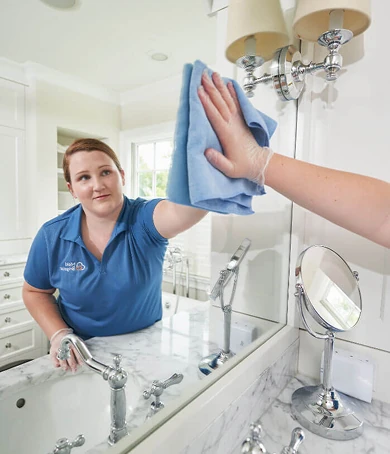 A person in a Maid Brigade uniform wipes a bathroom mirror with a cloth above a marble countertop and sink.